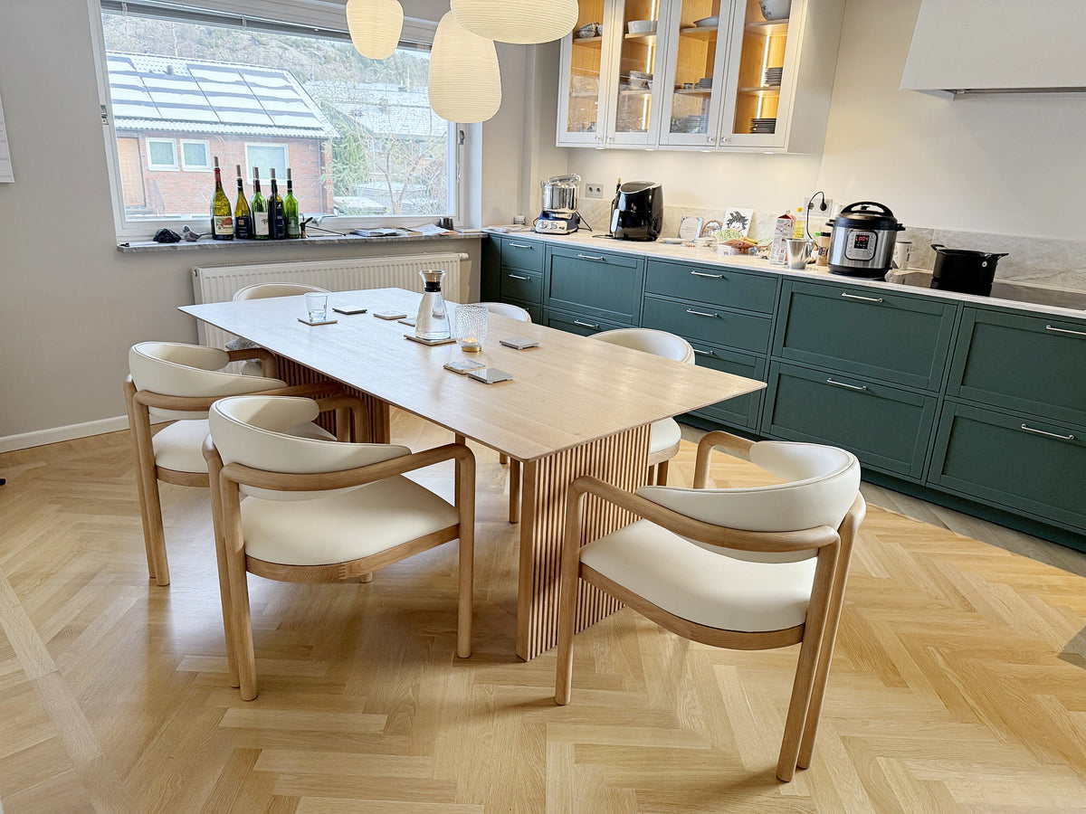 Dining area with rectangular wooden table surrounded by six light cushioned chairs, green cabinetry and kitchen appliances in background