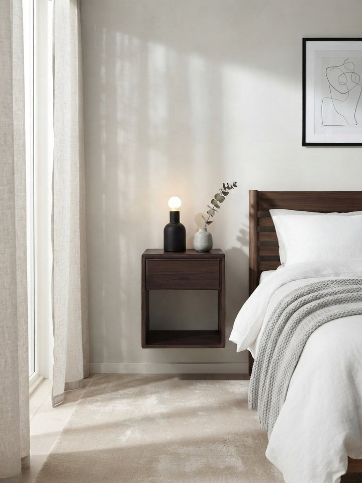 A modern bedroom scene featuring a dark brown floating nightstand beside a bed with white bedding and a gray knit throw. On the nightstand sits a black lamp and a small gray vase with dried stems.