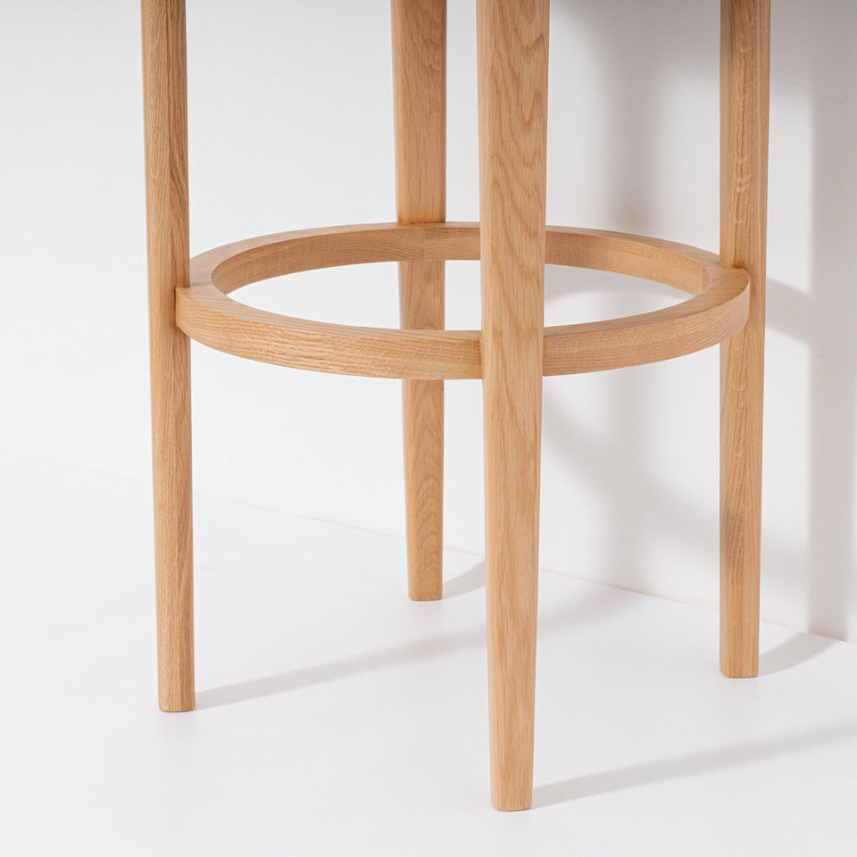 Close-up of a light natural oak wooden barstool showing the lower half with four legs and a circular footrest ring, set against a clean white background.