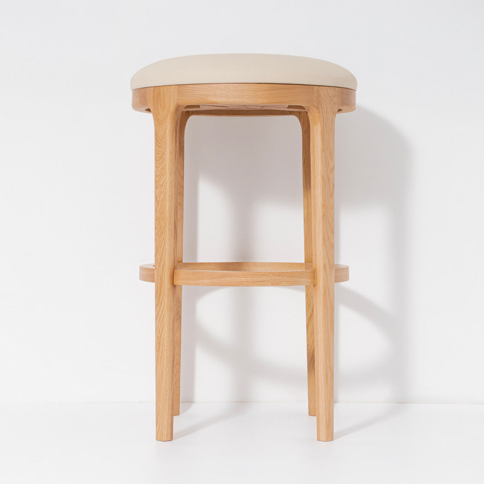A modern wooden bar stool with a light beige upholstered seat and a natural oak wood frame, set against a plain white background.