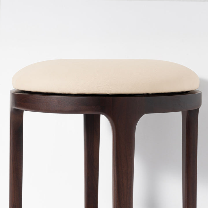 A close-up of a stool with a rounded, off-white upholstered seat cushion and a dark brown wooden frame and legs, set against a plain white background.