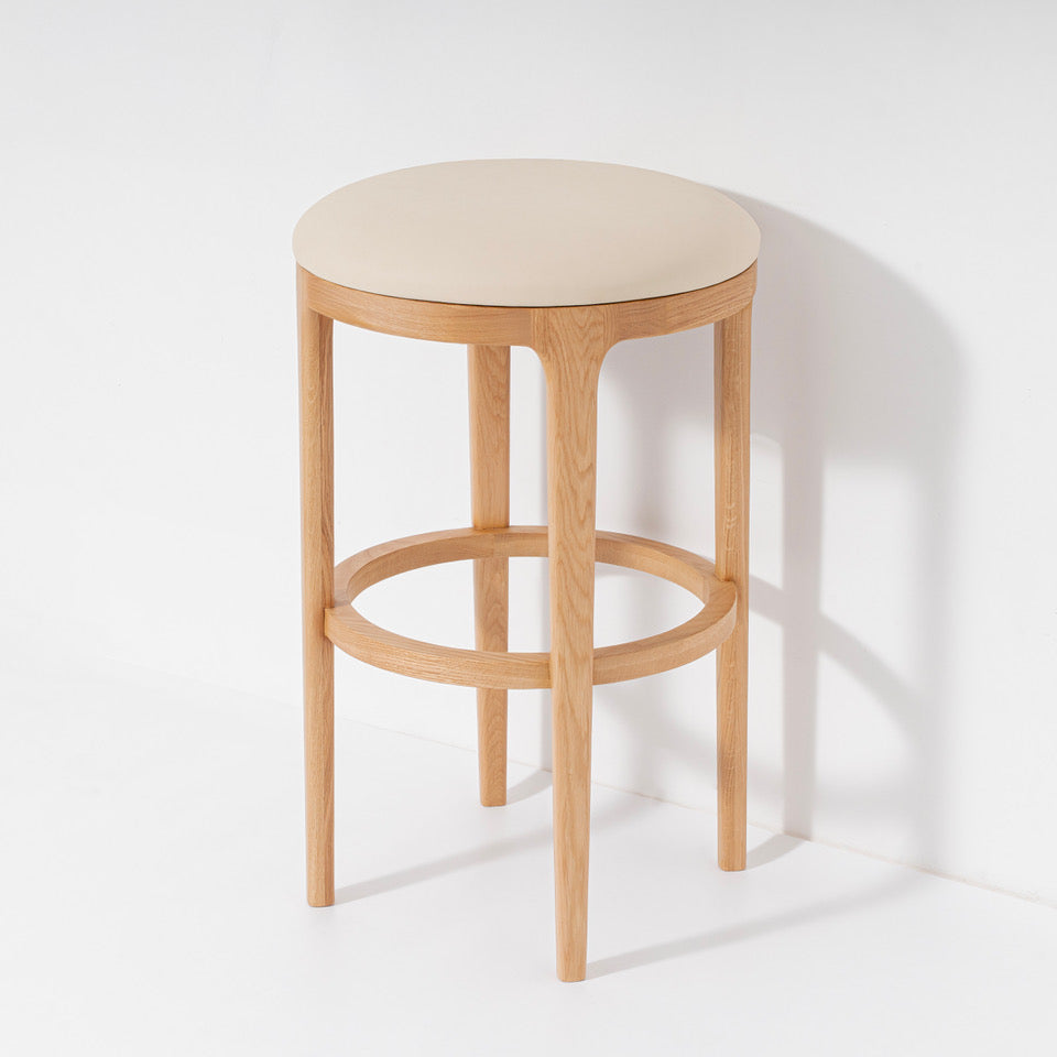 A modern wooden stool with a round light beige upholstered seat, four tapered natural oak legs, and a circular footrest, set against a clean white background with a soft shadow.
