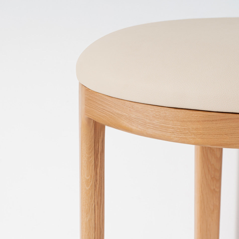 Close-up of a stool with a light beige upholstered seat and natural oak wood legs and rim, shown against a plain white background.