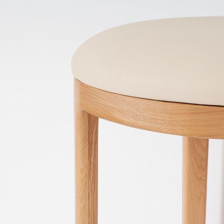 Close-up of a stool with a light beige upholstered seat and natural oak wood legs and rim, shown against a plain white background.