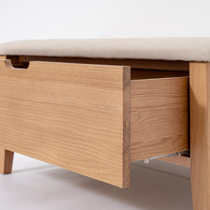 Close-up of a wooden storage bench or ottoman with a light beige upholstered top and an open drawer, showing the wood grain and interior.