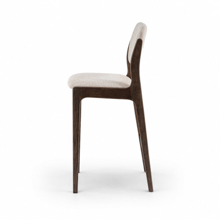 A modern barstool shown from the side against a solid white background, featuring a dark brown wood frame and light beige textured upholstery on the seat and backrest.