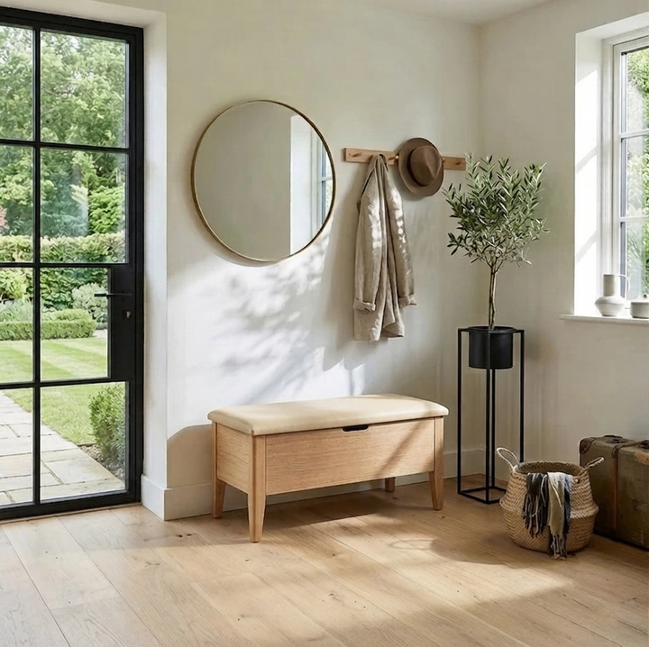 A light natural wood storage bench with a cream upholstered seat sits in a bright entryway. A round wall mirror, a wooden coat rack with a hat and jacket, a potted olive tree, and woven baskets are arranged around it.