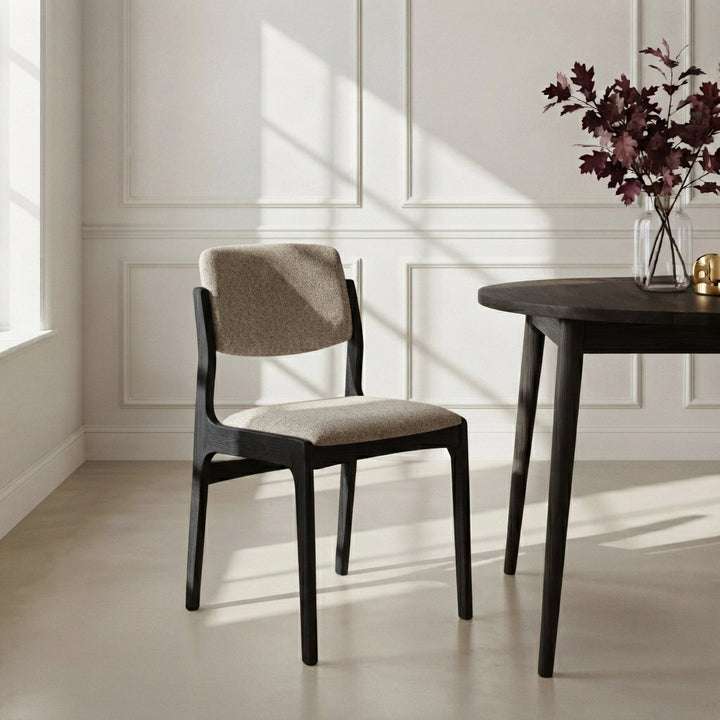 A modern dining chair with a black frame and textured beige upholstered seat and back, positioned next to a dark wood table in a bright room with sunlight and window panes.