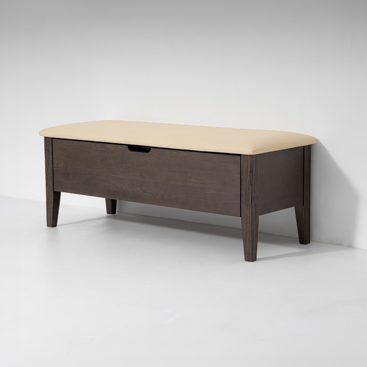 A modern storage bench with a dark brown wood frame, a single drawer, and a light beige upholstered top, shown against a plain light gray studio background.