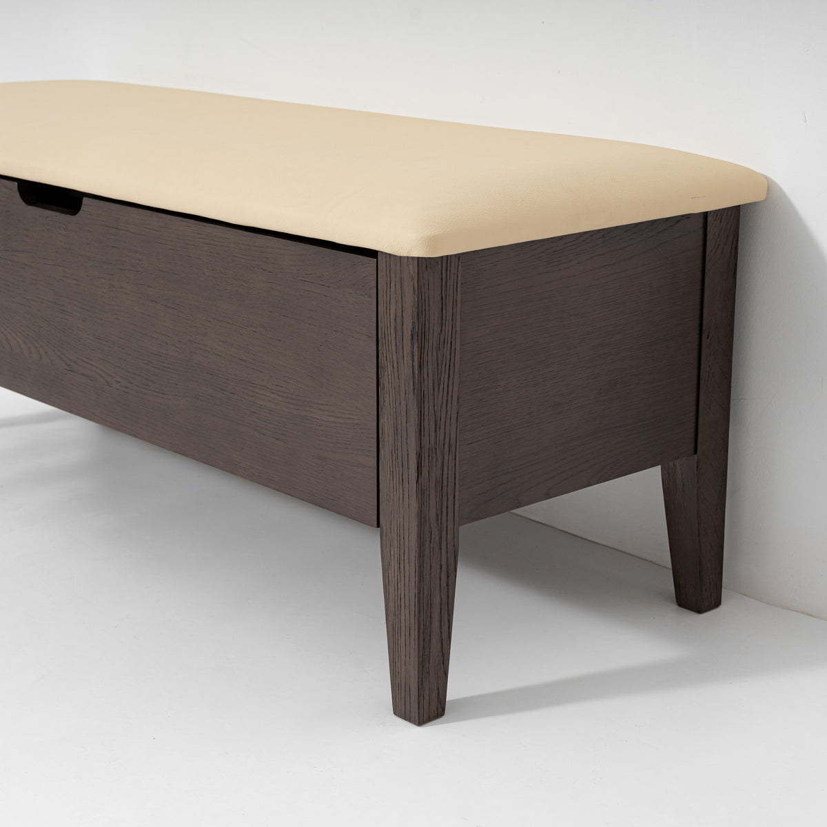 A modern wooden storage bench with a dark brown wood-grain frame and a light beige upholstered cushion on top, shown against a plain white wall.
