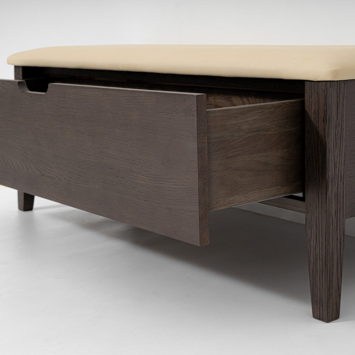 A modern wooden storage bench or coffee table with a dark brown wood finish, featuring a single open drawer and a light beige upholstered top cushion, shown on a light gray studio background.