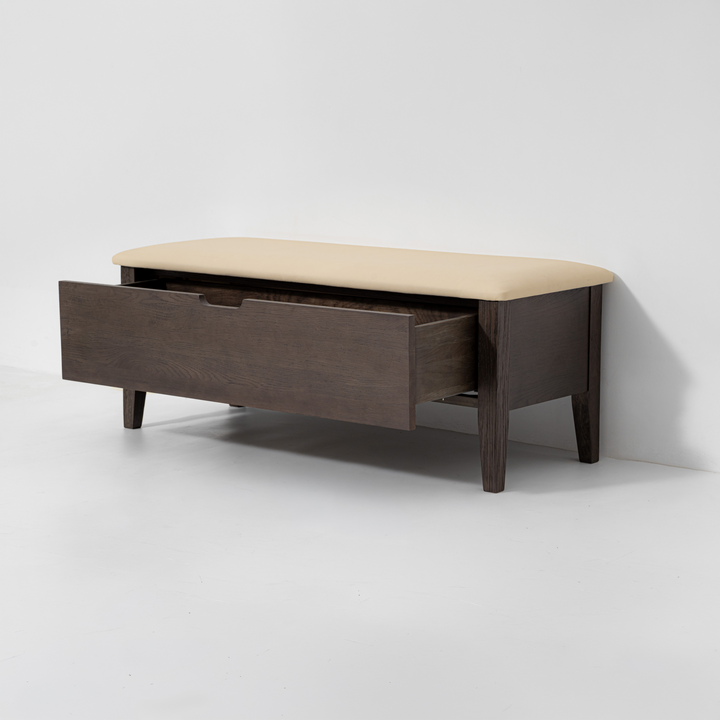A mid-century modern wooden storage bench with a dark brown wood frame, a single open drawer, and a beige upholstered cushion on top, set against a plain light gray wall and floor.
