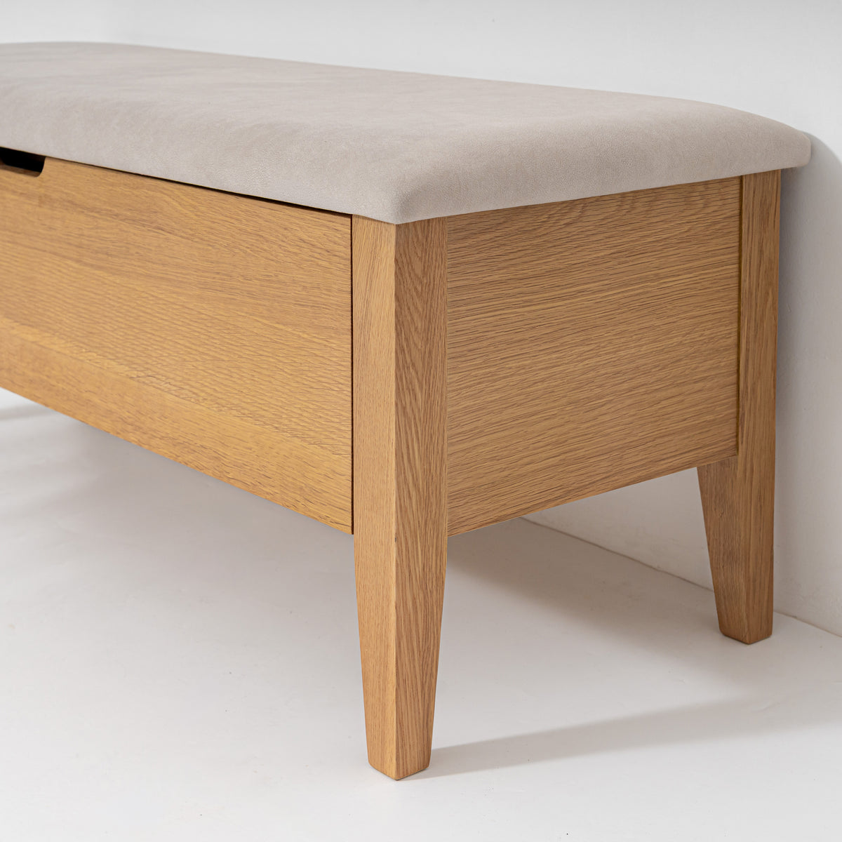 A close-up of a wooden storage bench with a light beige upholstered top and tapered legs, set against a plain white wall and floor.