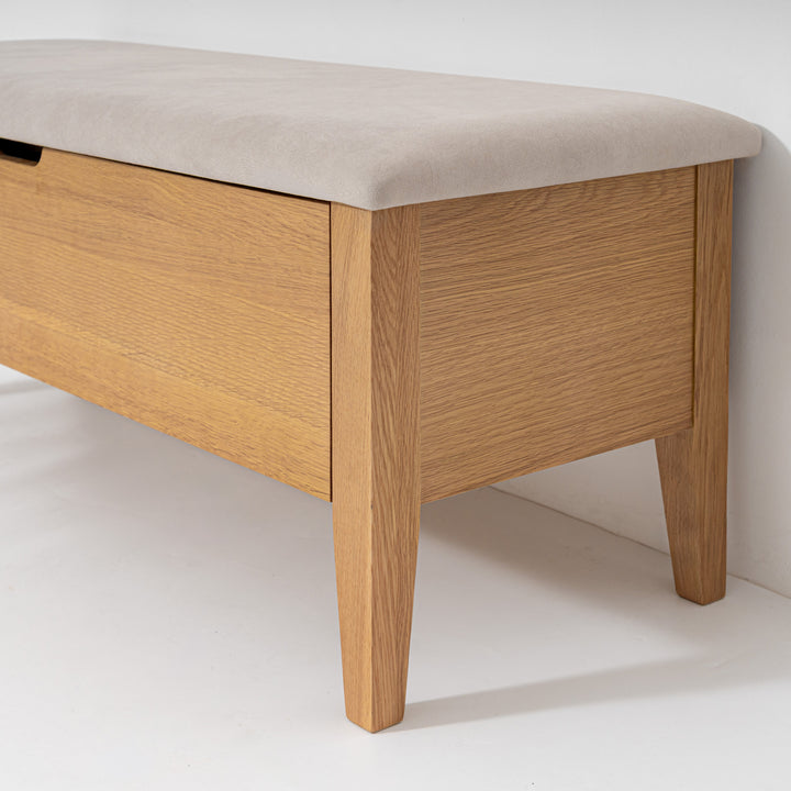 A close-up of a wooden storage bench with a light beige upholstered top and tapered legs, set against a plain white wall and floor.