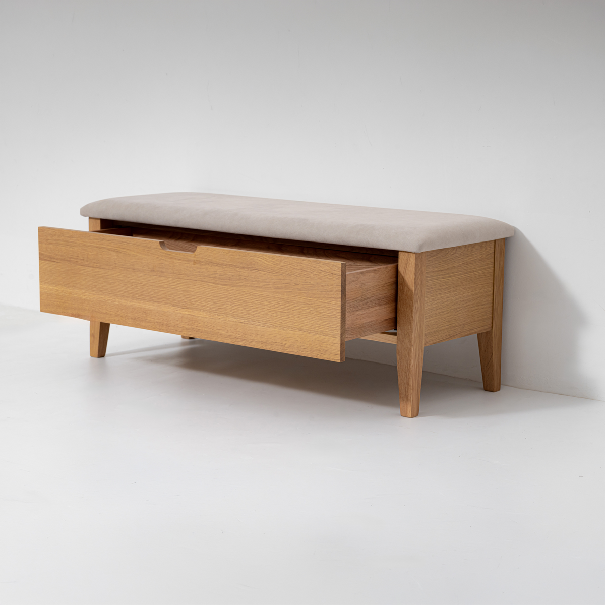 A mid-century modern wooden entryway bench with a light beige upholstered cushion on top and a single front drawer, shown against a plain light gray studio background.