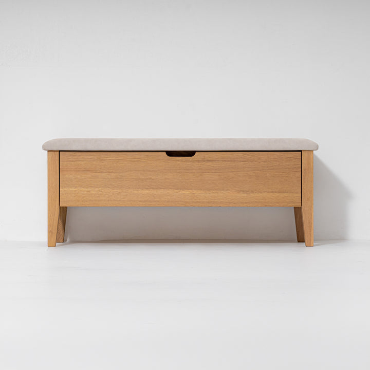 A mid-century modern wooden storage bench with a light oak wood frame, a single drawer with a dark handle, and a light beige upholstered top, shown against a plain white studio background.