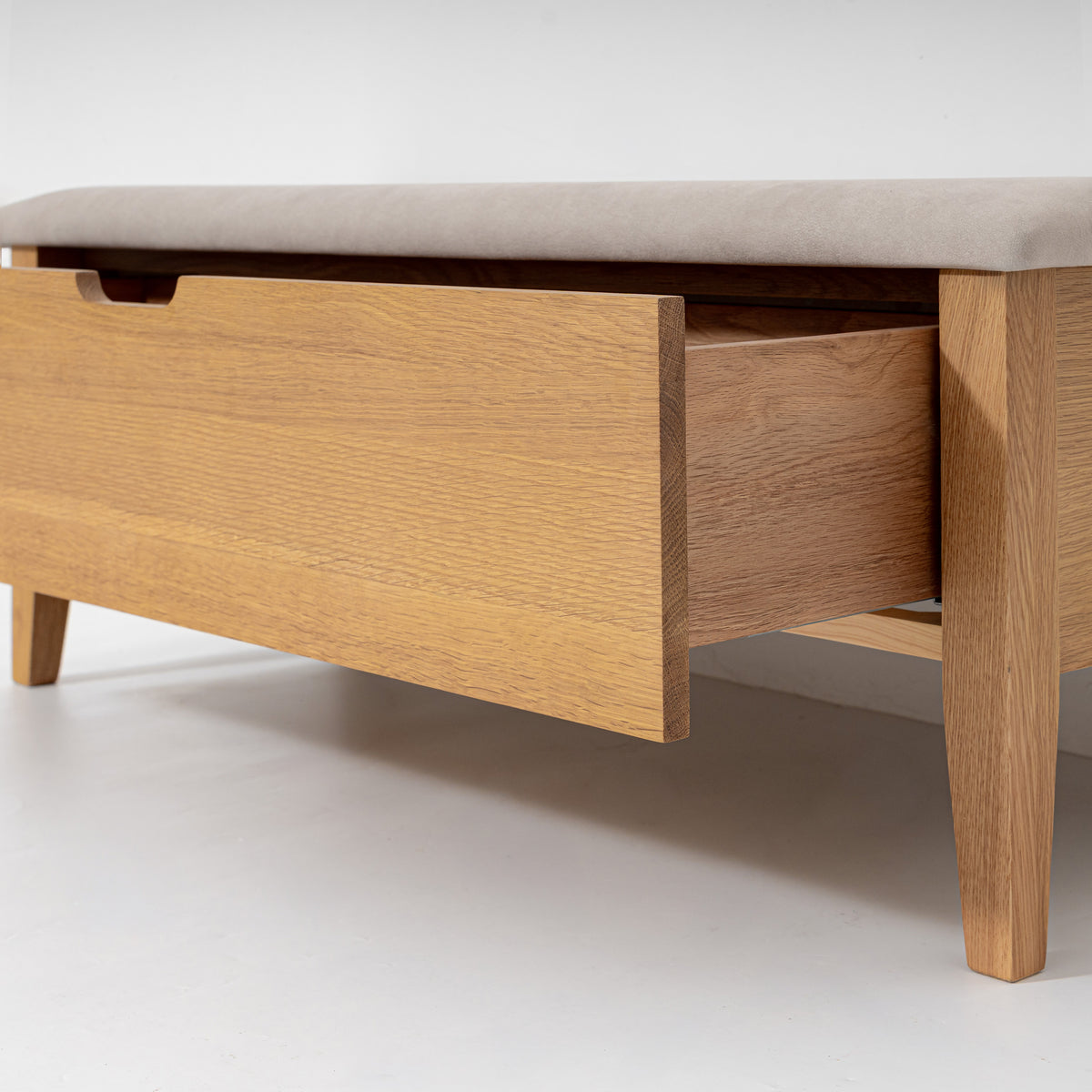 A close-up of a wooden storage bench or coffee table with a light beige upholstered top, one drawer pulled open, and tapered legs on a clean white studio background.