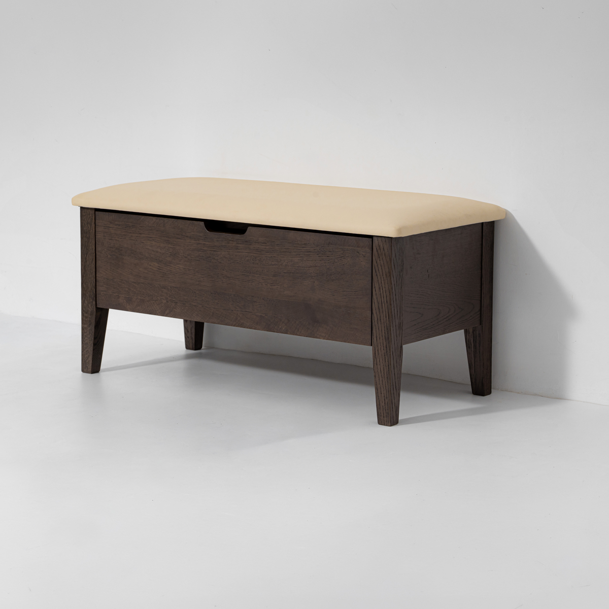 A modern wooden storage bench with a dark brown wood frame, a single drawer, and a light beige upholstered top, shown against a plain light gray wall and floor.