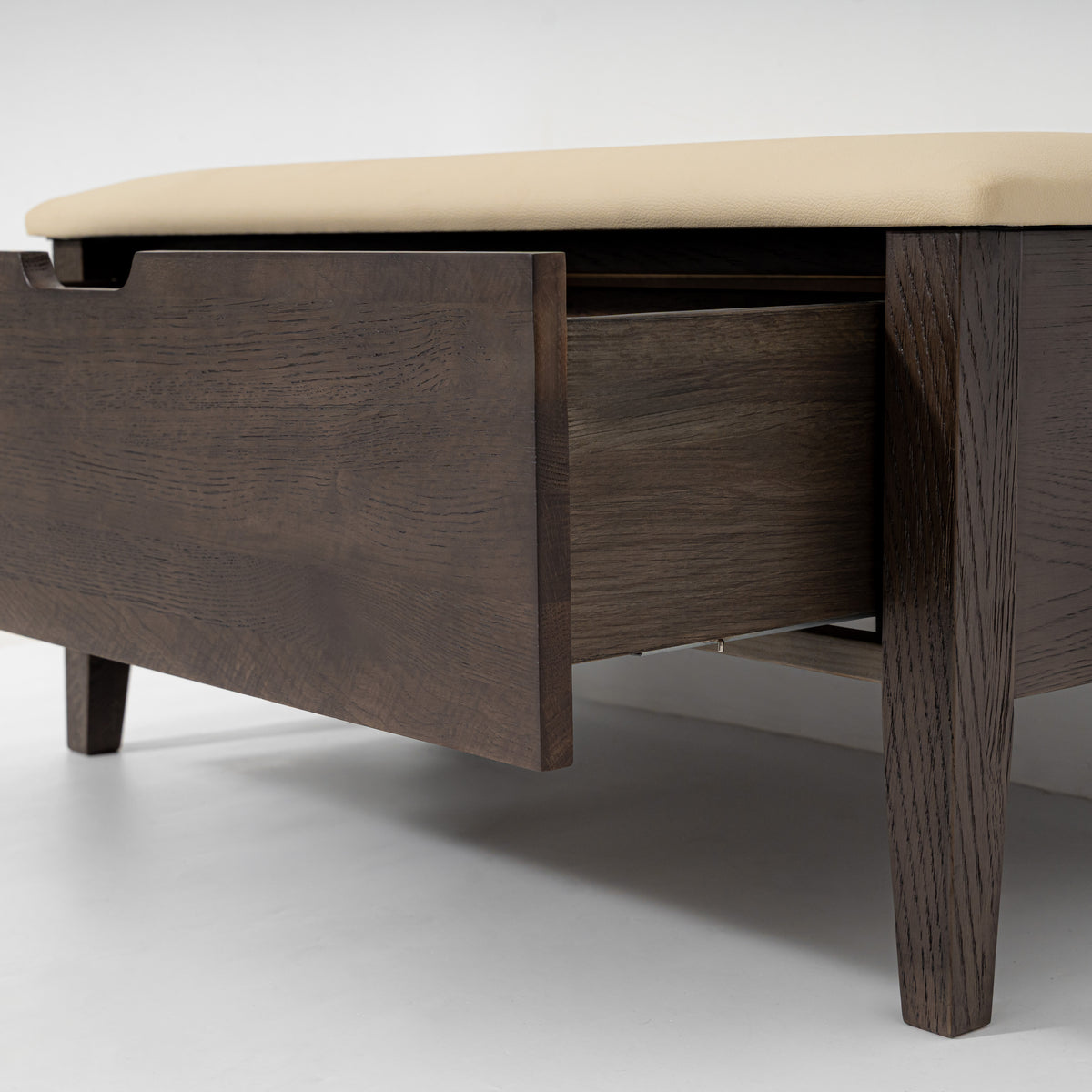 A close-up of a dark brown wooden storage bench or ottoman with a cream upholstered top and an open drawer, shown on a light gray studio background.