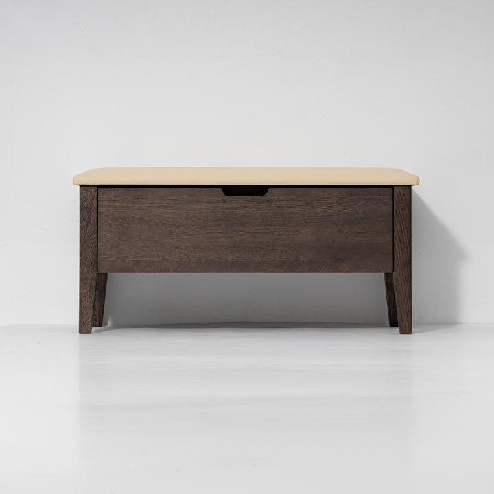 A modern storage bench with a dark brown wood frame, a single drawer with a black handle, and a light beige upholstered top, shown against a plain white studio background.