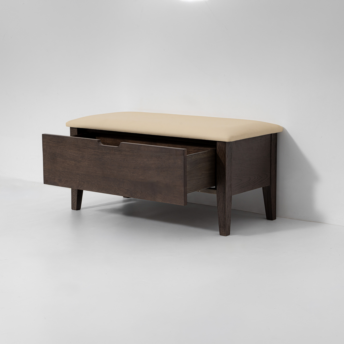 A dark brown wooden entryway bench with a beige upholstered seat cushion and one open drawer, shown against a plain light gray studio background.
