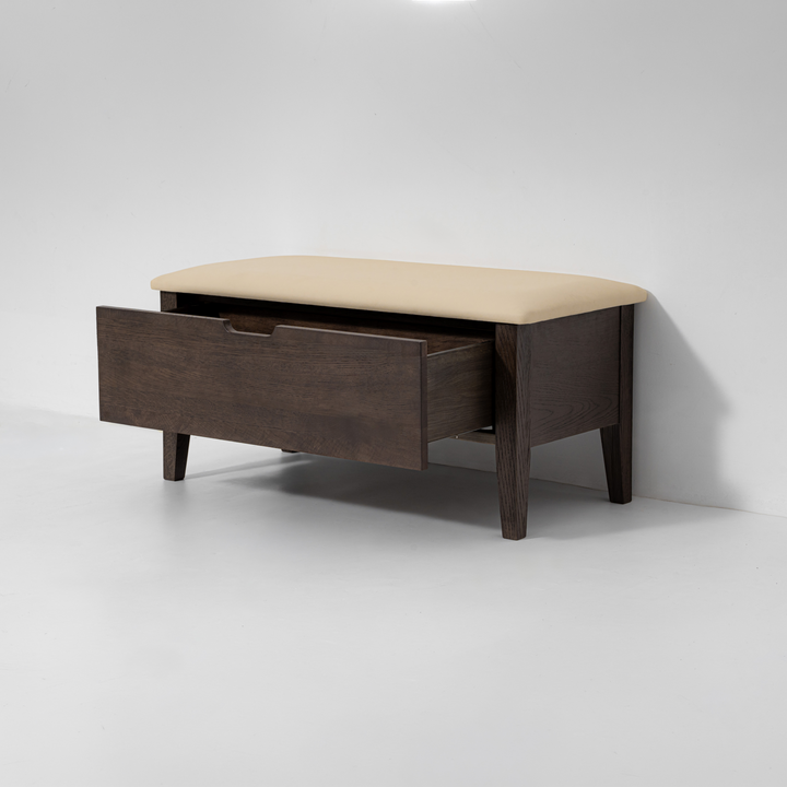 A dark brown wooden entryway bench with a beige upholstered seat cushion and one open drawer, shown against a plain light gray studio background.