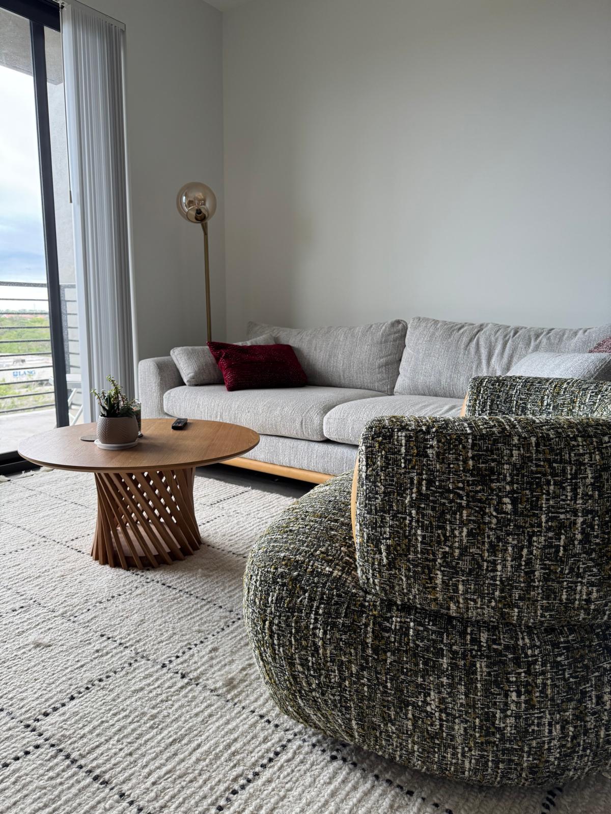 Living room with textured gray armchair, light gray sofa with red pillow, round wooden coffee table, floor lamp, and patterned area rug near sliding glass door.