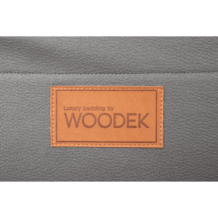 A brown leather patch reading "Luxury bedding by WOODEK" is stitched onto the grey fabric surface of the Woodek 2-in-1 Dog Travel Bag & Mattress, ideal as a portable dog bed for travel or home use.